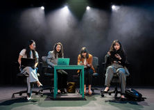 4 People sat in office chairs around a table on stage lit.