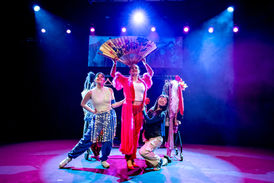 3 people stood centrally on stage lit, a clothes rail is in the background.